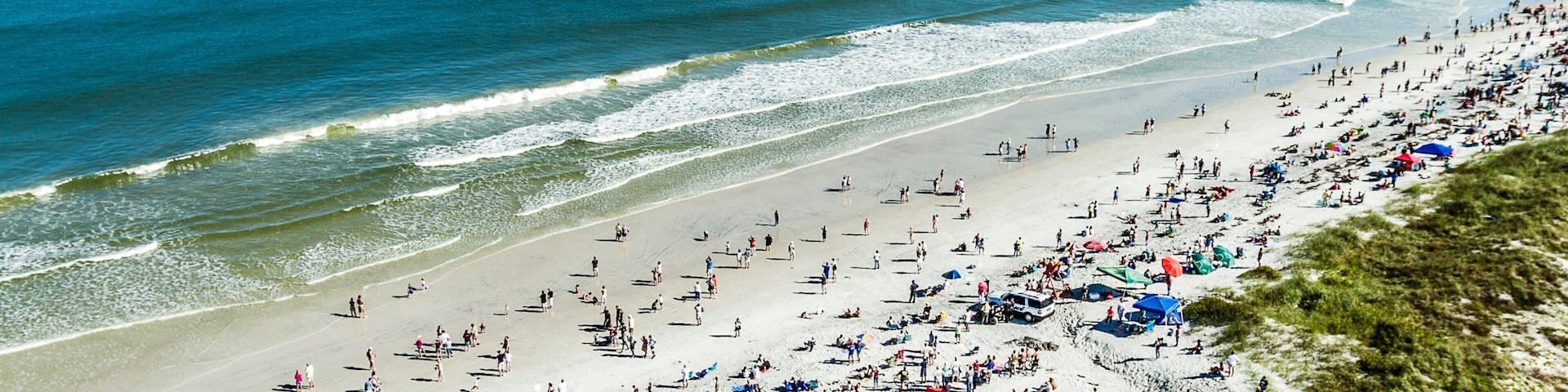 A crowded beach with people, umbrellas, and a pier in the background.