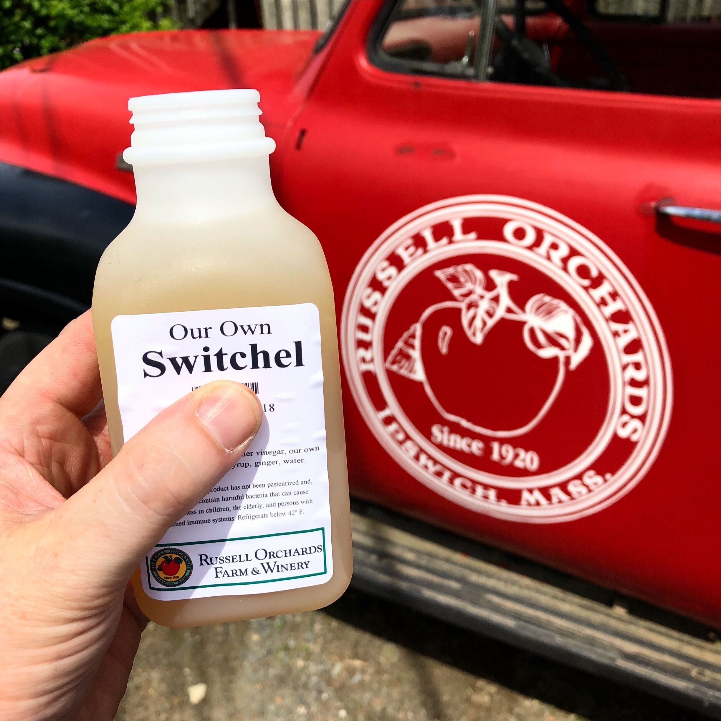 A hand holding a bottle of 'Our Own Switchel' vinegar with a label from Russell Orchards Farm & Winery, next to a red vehicle with the same branding.