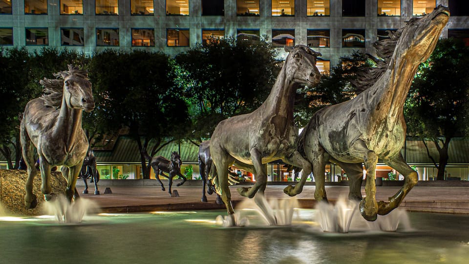 A nighttime sculpture of horses in motion, set against a backdrop of illuminated office buildings.