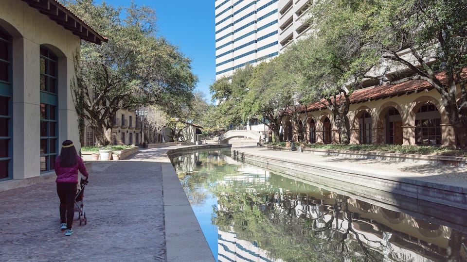 A canal with a person pushing a stroller, surrounded by trees and historic buildings.