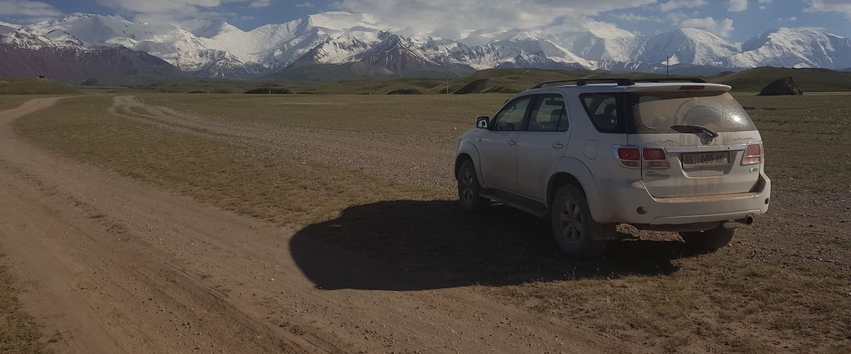 A white SUV parked on a dirt road with snow-capped mountains in the background.