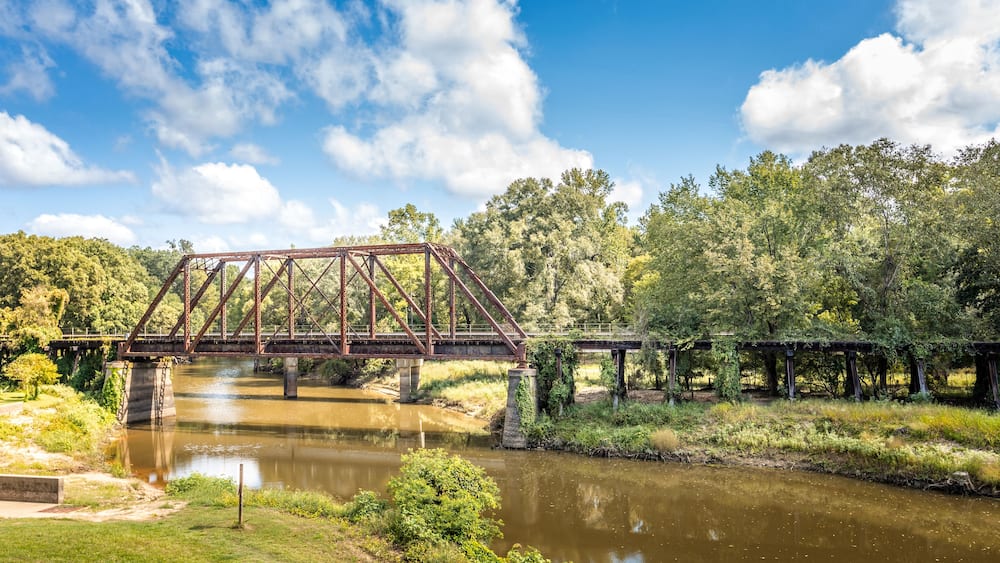 Eine verrostete Metallbrücke über einem ruhigen Fluss, umgeben von üppigem Grün und Bäumen.