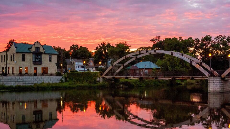 A bridge over a calm river at sunset with a reflection of the sky and buildings.