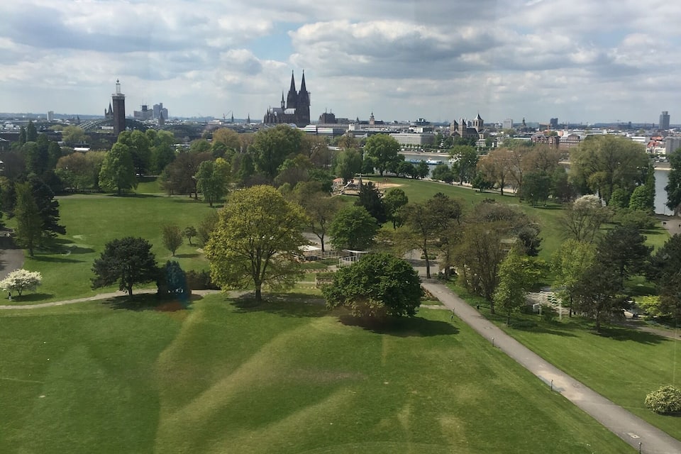 Ein Park mit einer großen, grünen Wiese, Bäumen und einem gewundenen Weg. Im Hintergrund eine Stadtansicht mit verschiedenen Gebäuden und einer markanten Kirche.