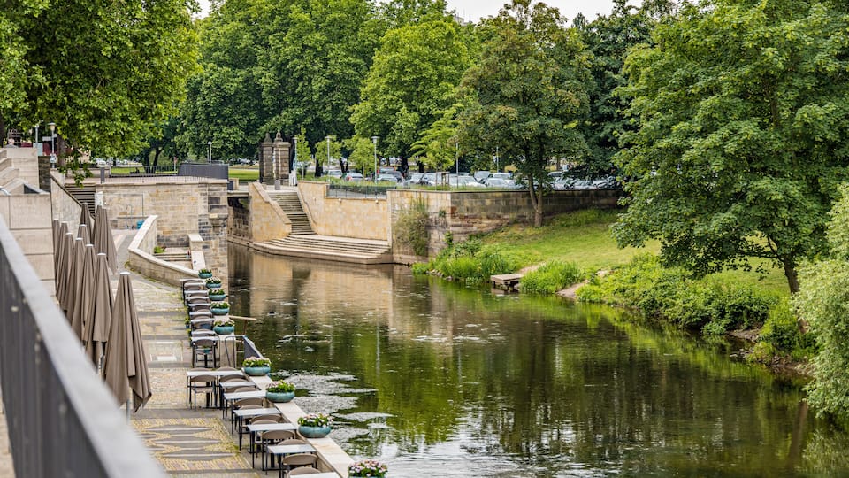 A riverside café with outdoor seating overlooking a calm river, surrounded by lush greenery and stone structures.