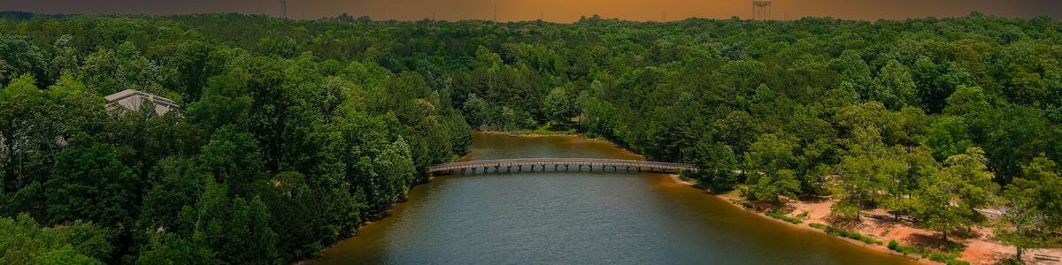 a gorgeous aerial shot of the rippling lake water surrounded by lush green trees, grass and plants with a gorgeous powerful clouds at sunset and a brown wooden bridge over the water at Indian Lake