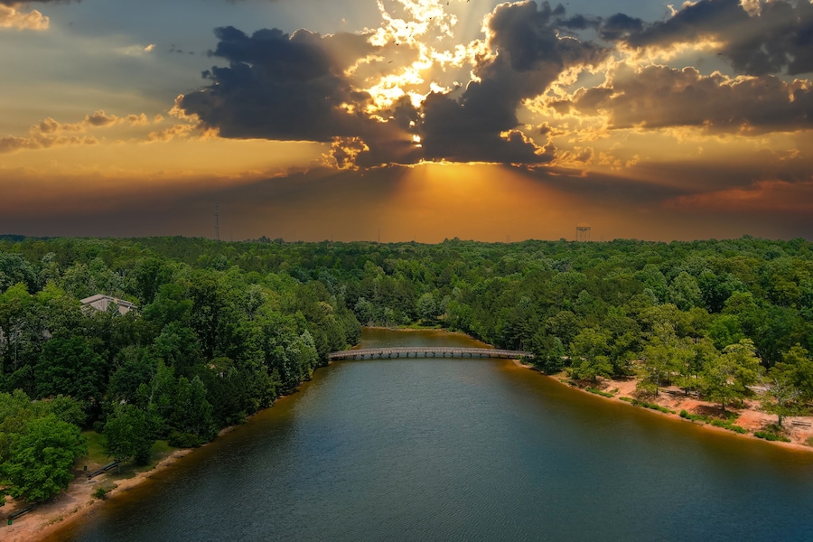 a gorgeous aerial shot of the rippling lake water surrounded by lush green trees, grass and plants with a gorgeous powerful clouds at sunset and a brown wooden bridge over the water at Indian Lake