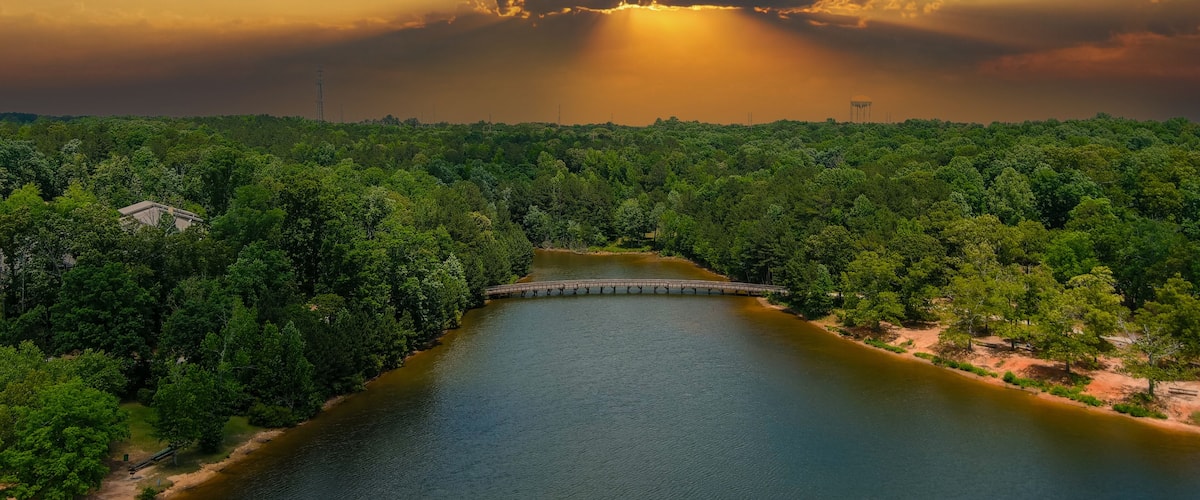 a gorgeous aerial shot of the rippling lake water surrounded by lush green trees, grass and plants with a gorgeous powerful clouds at sunset and a brown wooden bridge over the water at Indian Lake