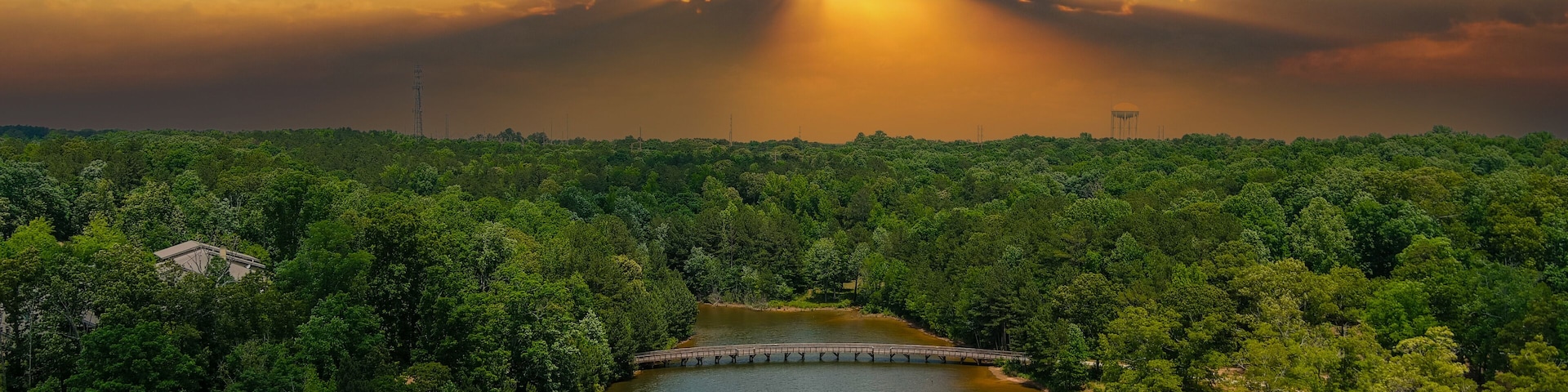 a gorgeous aerial shot of the rippling lake water surrounded by lush green trees, grass and plants with a gorgeous powerful clouds at sunset and a brown wooden bridge over the water at Indian Lake