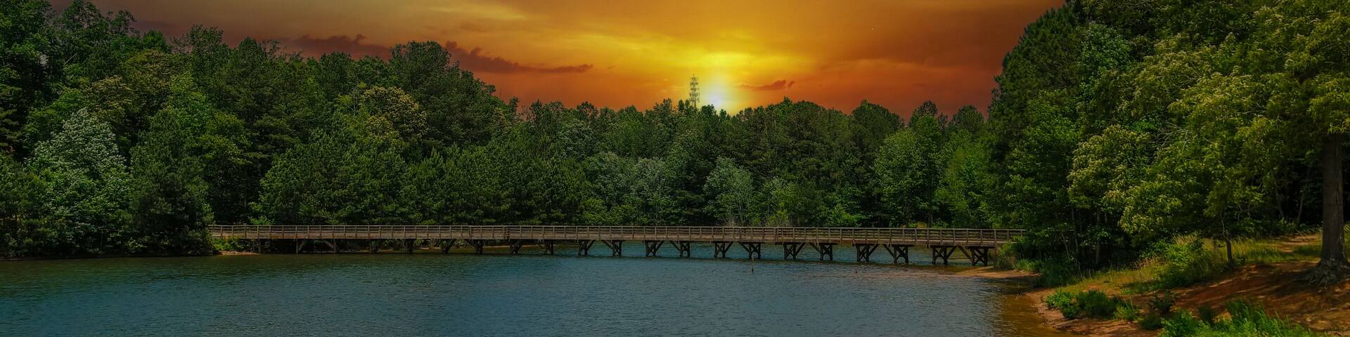 a gorgeous aerial shot of the rippling lake water surrounded by lush green trees, grass and plants with a gorgeous powerful clouds at sunset and a brown wooden bridge over the water at Indian Lake