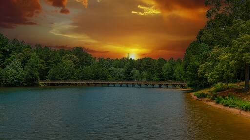 a gorgeous aerial shot of the rippling lake water surrounded by lush green trees, grass and plants with a gorgeous powerful clouds at sunset and a brown wooden bridge over the water at Indian Lake