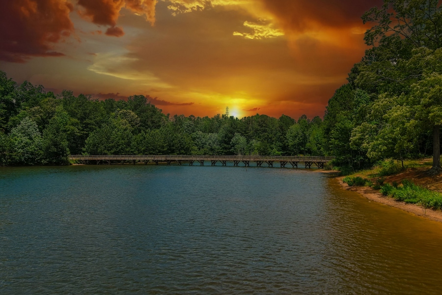 a gorgeous aerial shot of the rippling lake water surrounded by lush green trees, grass and plants with a gorgeous powerful clouds at sunset and a brown wooden bridge over the water at Indian Lake