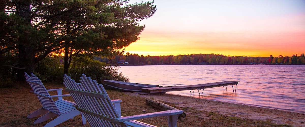 Summer Vacation At The Lake. Sunset over a lake with a row of Adirondack Chairs, dock and aluminum rowboat.