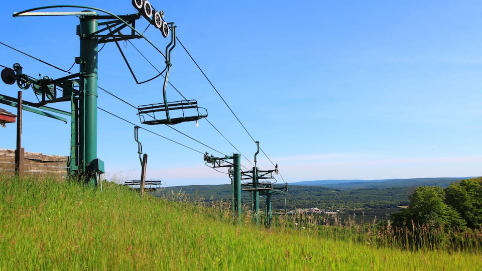 A ski lift with cables and chairs against a clear blue sky, surrounded by lush green grass and distant hills.