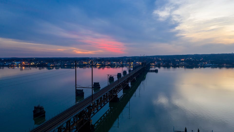 A calm river with a long bridge extending into the distance, surrounded by a cityscape with lights reflecting on the water.