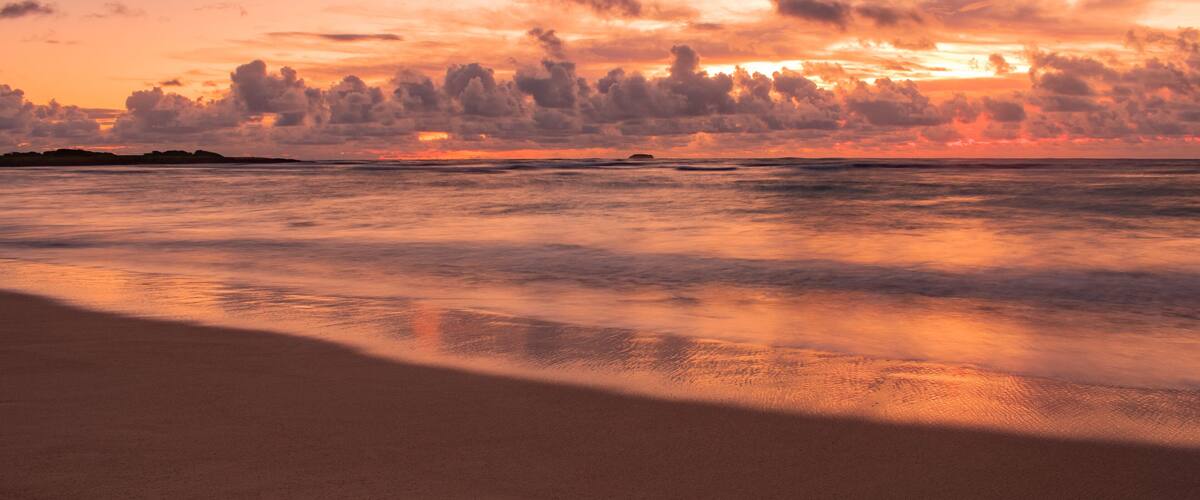 A beach at sunset with a calm sea and a sky filled with clouds.