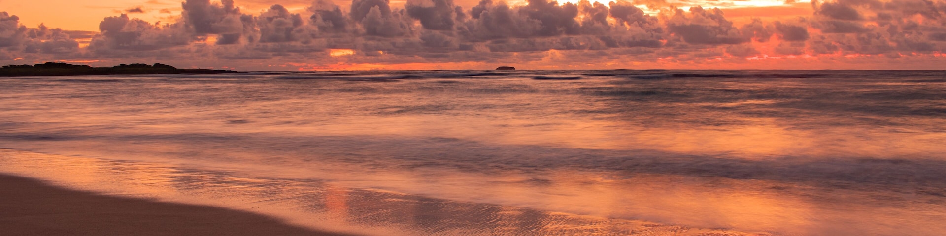 A beach at sunset with a calm sea and a sky filled with clouds.