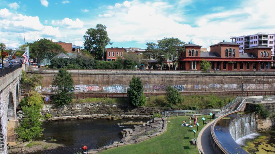 A river with a stone bridge, a park with people, and buildings in the background.