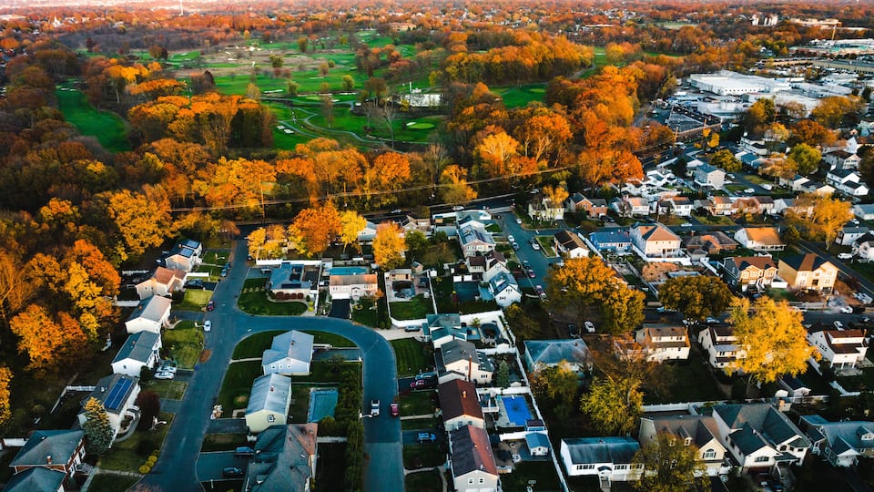 An aerial view of a residential area with houses, roads, and autumn-colored trees.
