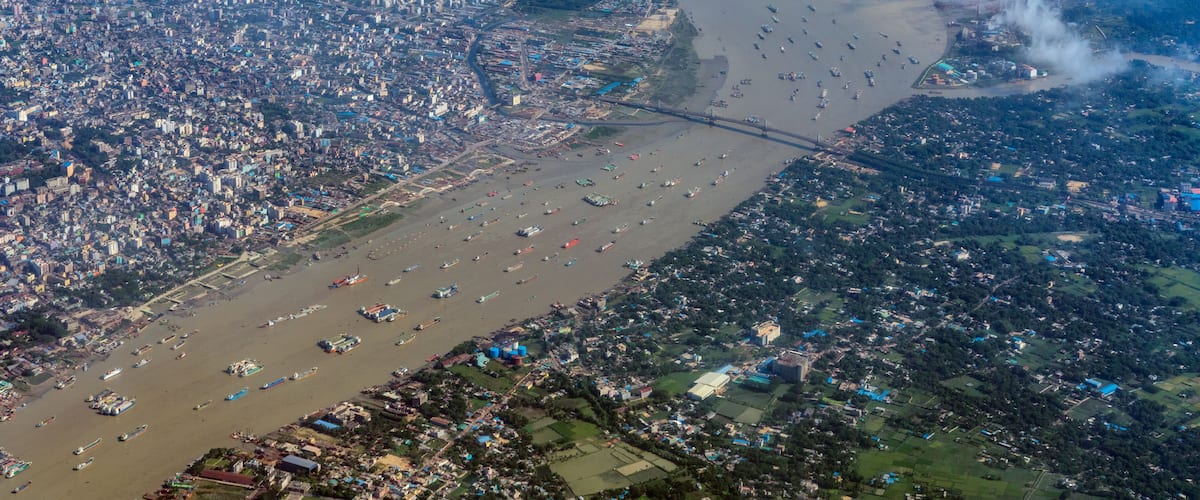 An aerial view of a river with numerous boats, a bridge, and densely packed urban and rural areas.