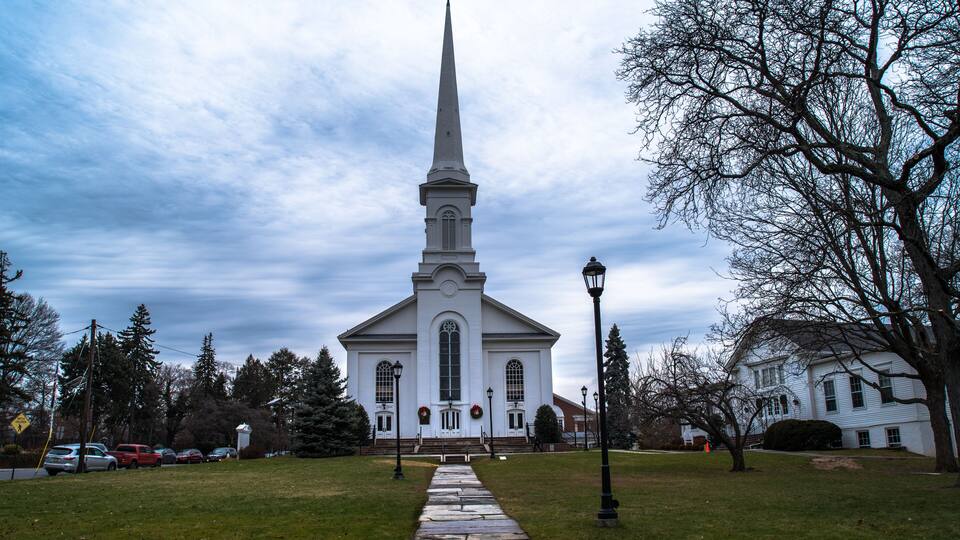 A white church with a tall steeple, a stone pathway leading to the entrance, and surrounding trees and houses.