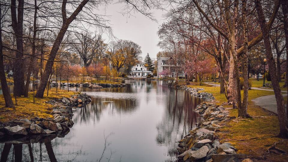 A serene park with a pond, autumn-colored trees, and a white house in the background.