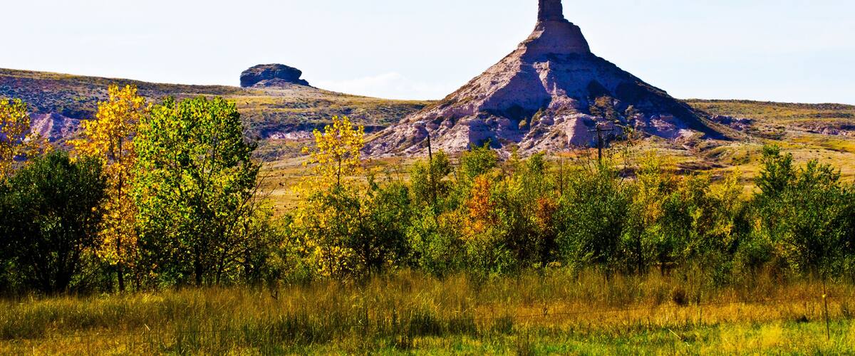 A prominent butte with a pointed peak, surrounded by autumn-colored trees and a clear sky.
