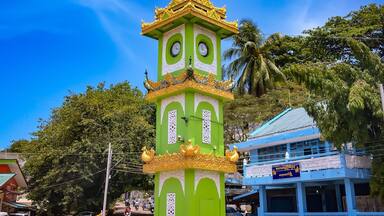 A green and gold tower with clocks, surrounded by palm trees and a blue building.
