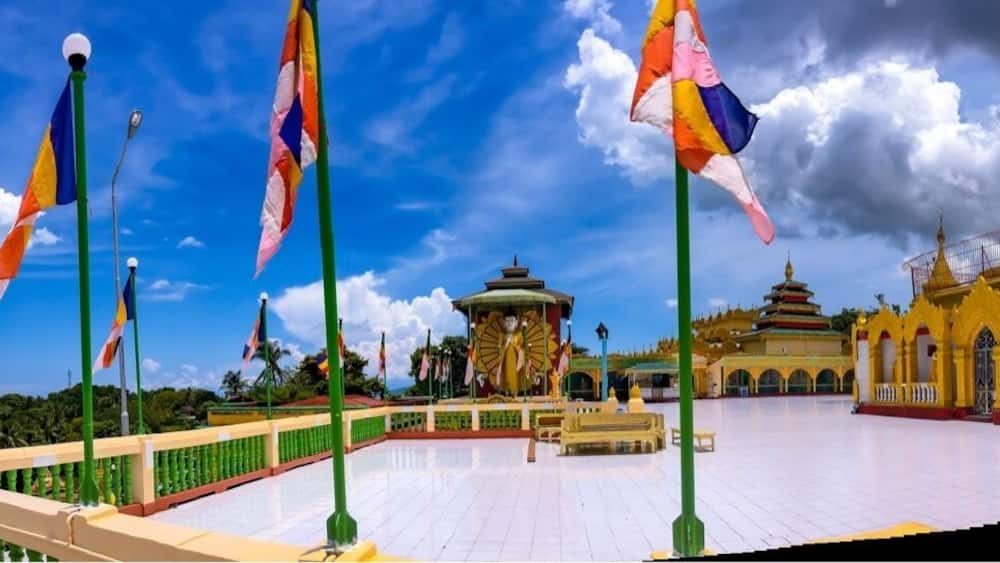 A golden pagoda, a green-roofed pavilion, and colorful flags against a blue sky.