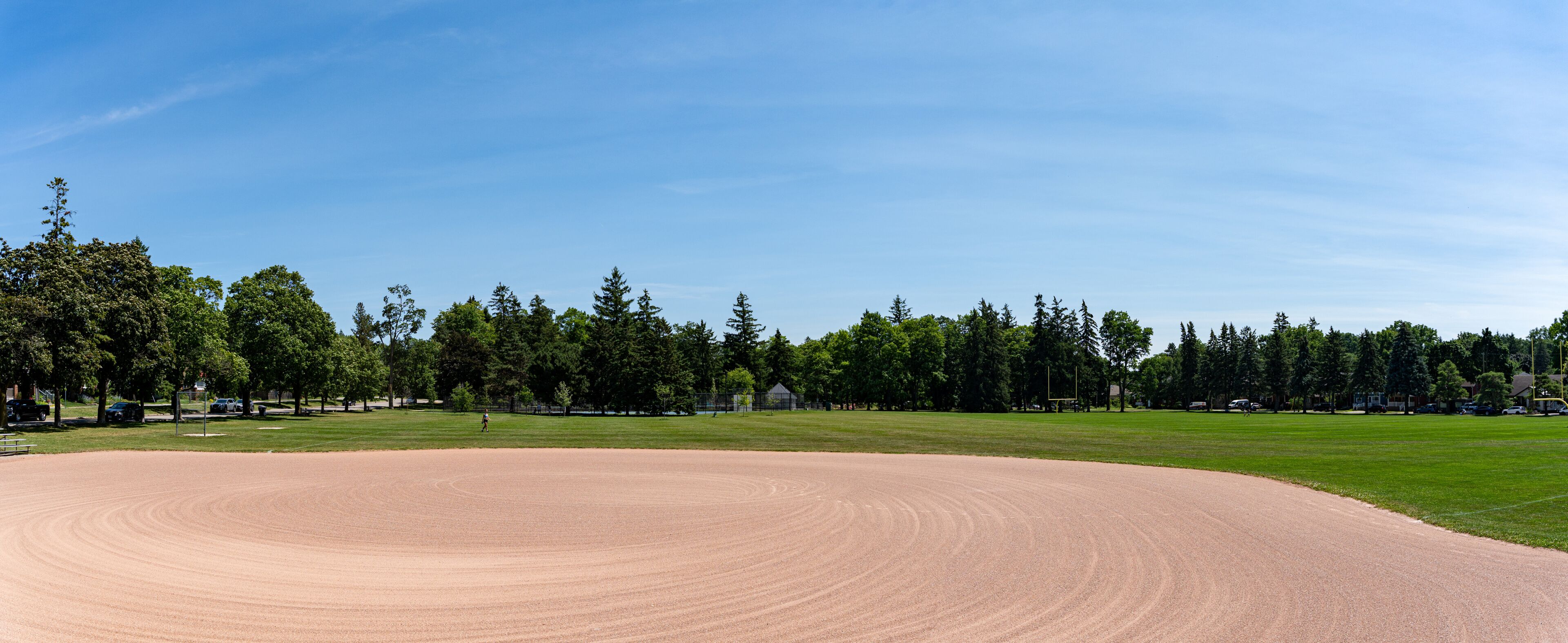 En baseballbane med jorddækket infield og græsbelagt outfield, omgivet af træer og en klar blå himmel.