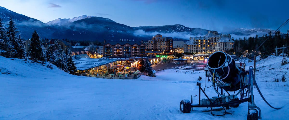 Un complejo turístico en la montaña cubierto de nieve al anochecer, con edificios iluminados y un telesilla.