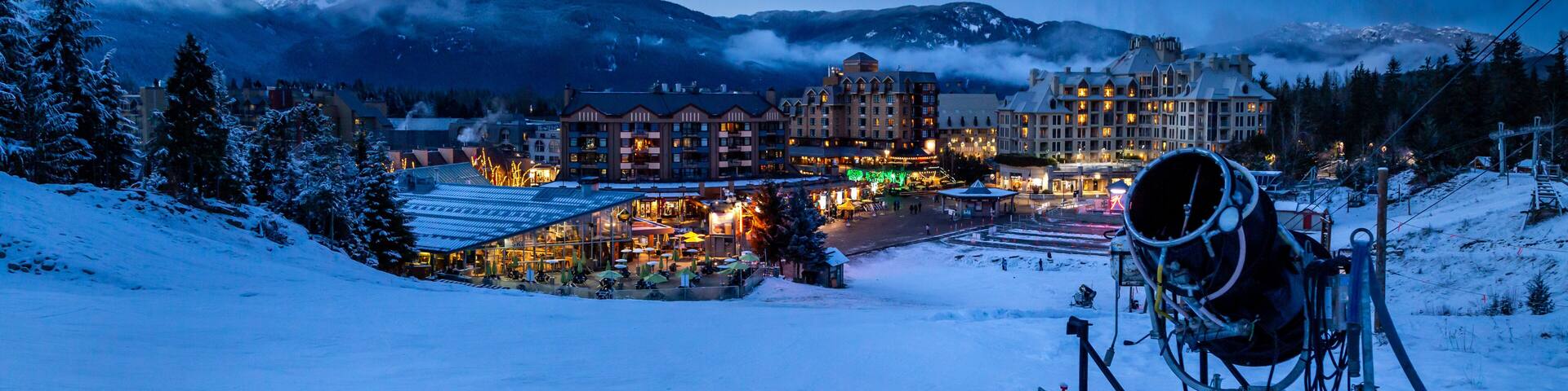 A snow-covered mountain resort at dusk with illuminated buildings and a ski lift.