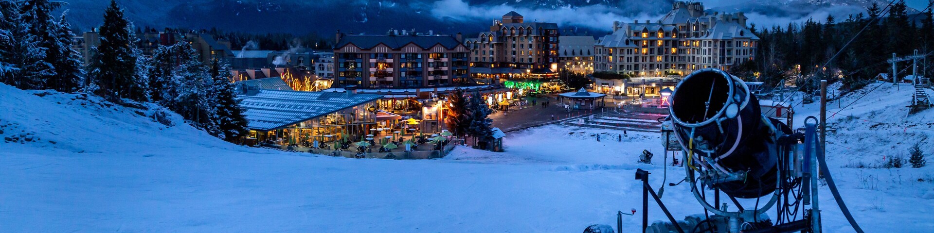A snow-covered mountain resort at dusk with illuminated buildings and a ski lift.