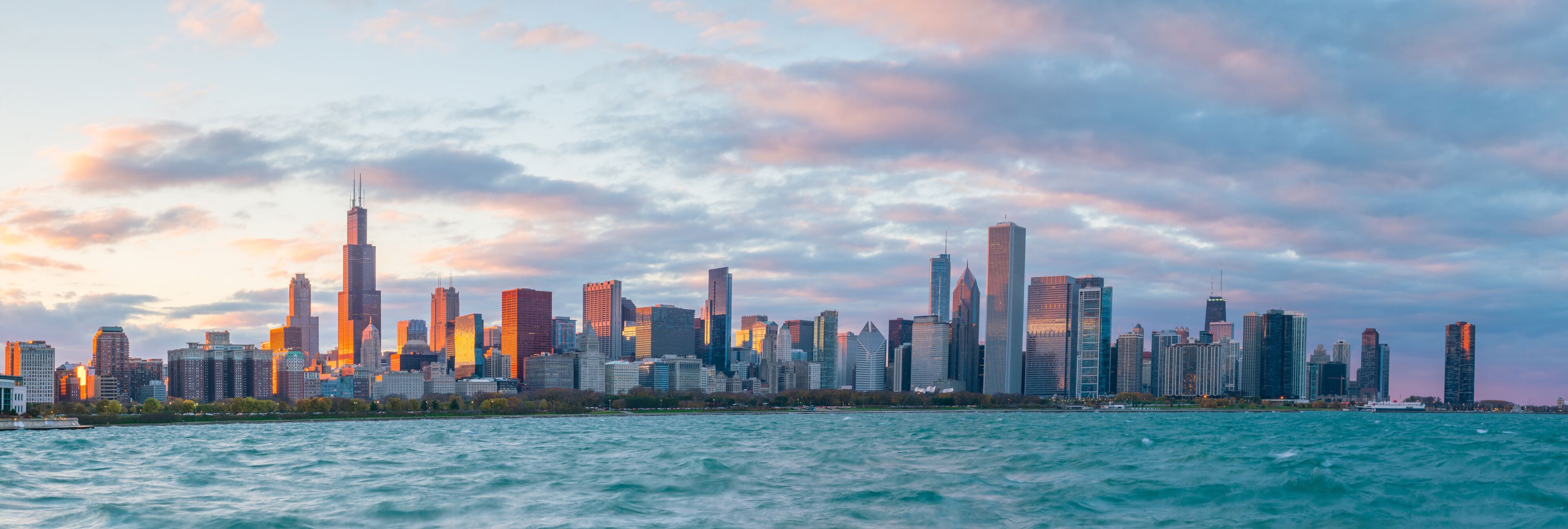 A city skyline with numerous high-rise buildings, a body of water in the foreground, and a sunset sky.