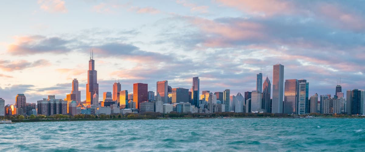 A city skyline with numerous high-rise buildings, a body of water in the foreground, and a sunset sky.