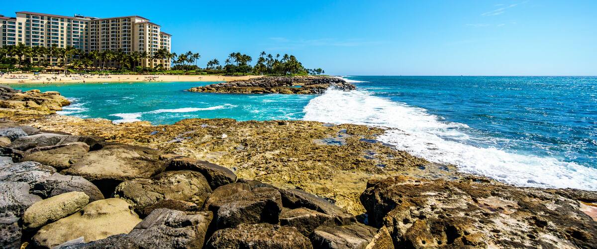 A coastal scene with rocky shores, clear blue waters, a sandy beach, and a hotel complex in the background.
