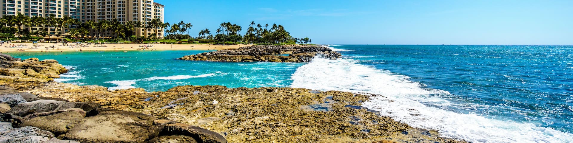 A coastal scene with rocky shores, clear blue waters, a sandy beach, and a hotel complex in the background.