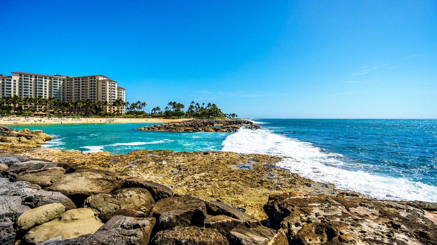 A coastal scene with rocky shores, clear blue waters, a sandy beach, and a hotel complex in the background.