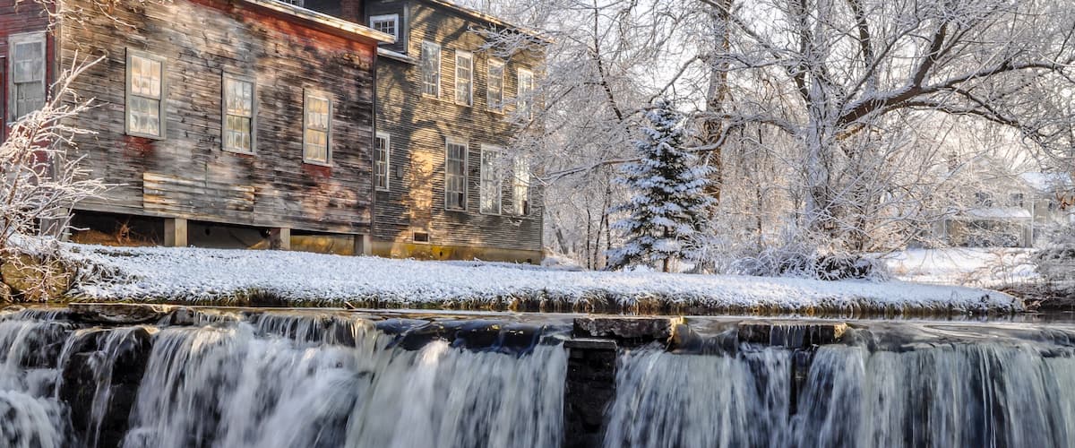 A waterfall in a snowy landscape with a wooden building in the background.
