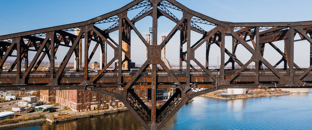 A large steel bridge with a triangular truss design spanning over a body of water, with industrial buildings and a clear sky in the background.