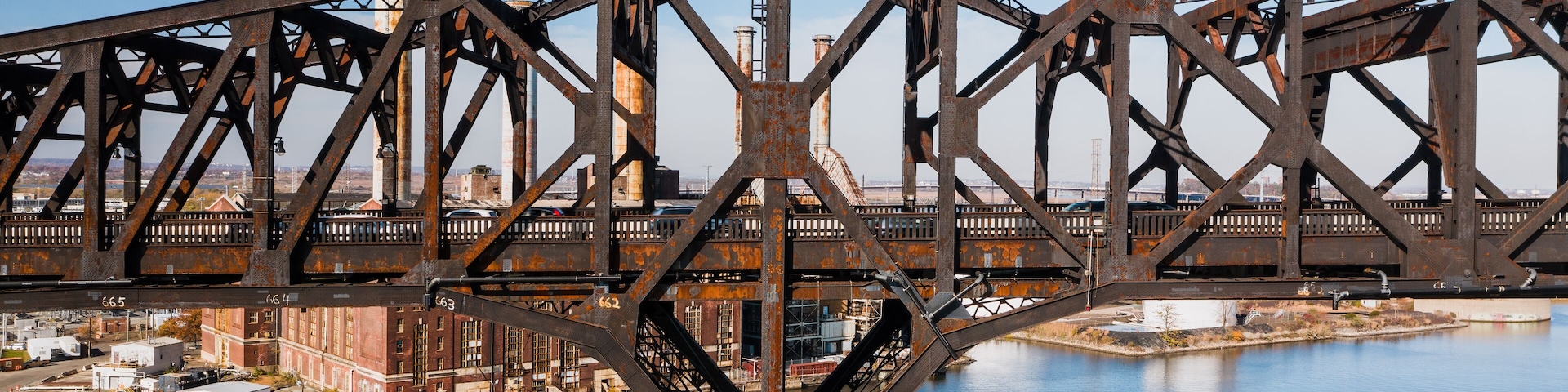 A large steel bridge with a triangular truss design spanning over a body of water, with industrial buildings and a clear sky in the background.
