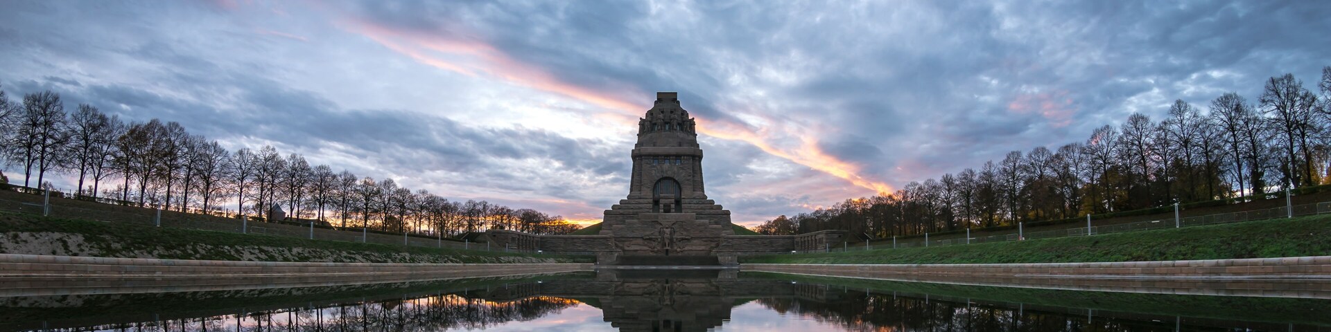 Um monumento de pedra refletido em águas calmas, rodeado por árvores e um céu nublado.