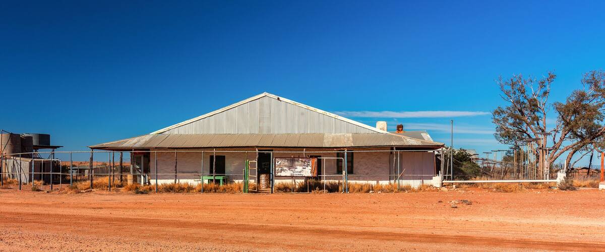 A rural house with a corrugated metal roof, surrounded by dry grass and a dirt road.