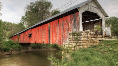 A red covered bridge with a stone foundation, surrounded by greenery and a river.