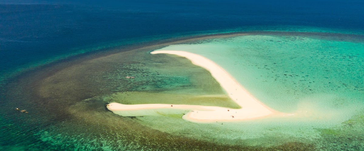 A clear aerial view of a coastal landscape with a prominent white sandy beach, a narrow water channel, and a backdrop of distant landforms.
