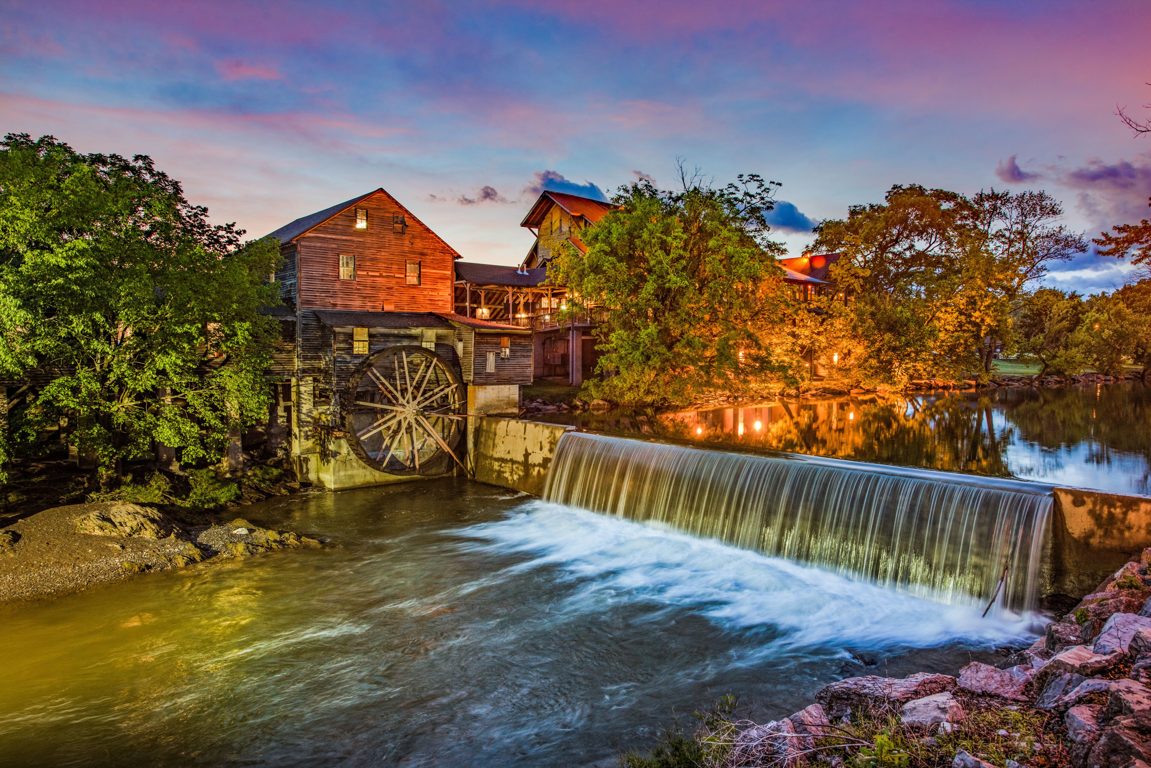 A water mill with a wooden structure and a large wheel, surrounded by trees and a calm river.