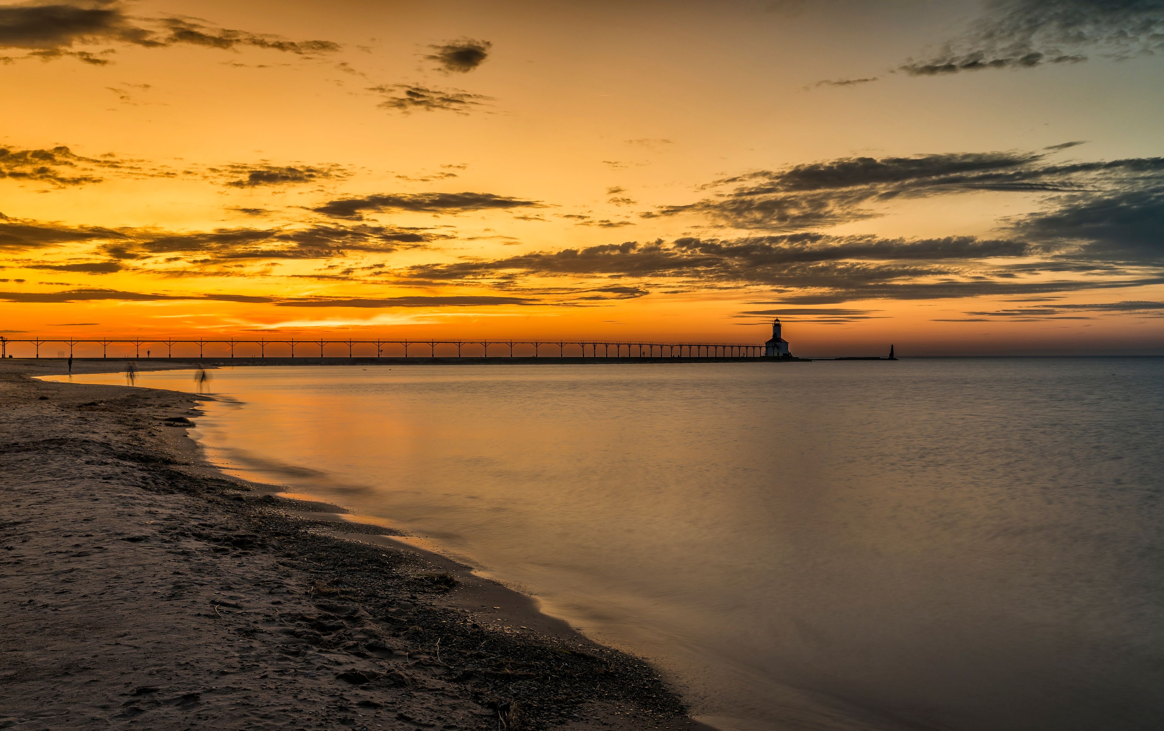 A sunset over a calm sea with a pier extending into the water, a lighthouse, and a sandy beach.
