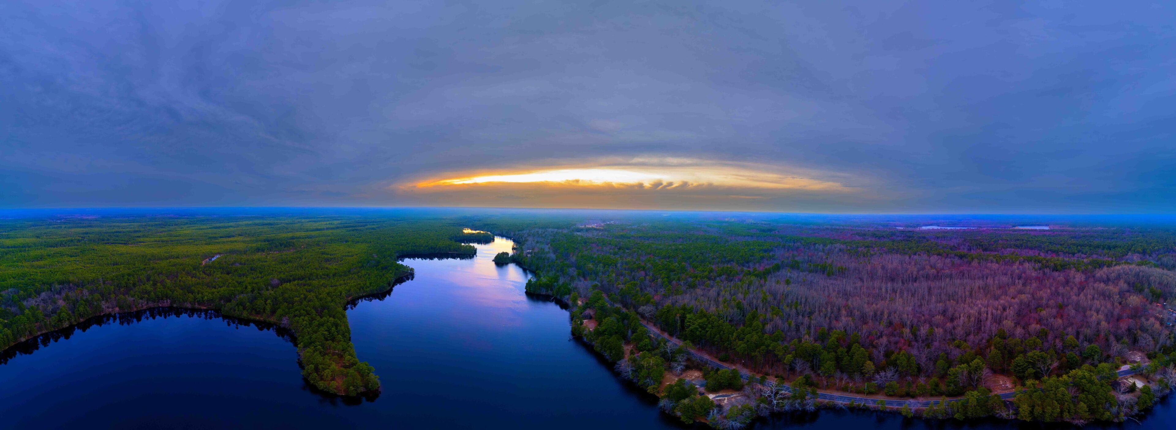 A river winding through a forested landscape with a sunset in the distance.