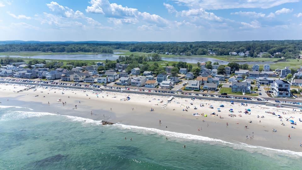 A coastal town with a sandy beach, a promenade lined with houses, and a backdrop of hills and greenery.