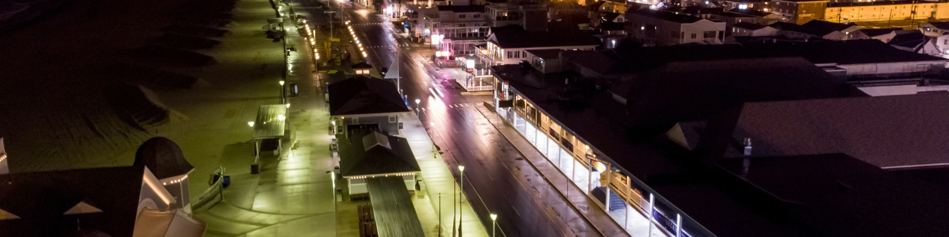 Vista aérea nocturna de un pueblo costero con edificios iluminados, un paseo marítimo y una estación de tren.